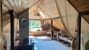 An attic room with a wooden ceiling, a bed, a reading chair, a small table, and a window with a view of greenery.