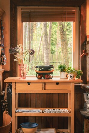 Una mesa de madera con vista a árboles, un jarro con flores, una cesta y una planta en maceta.