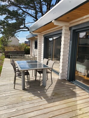Une terrasse extérieure moderne, dotée d’une table et de chaises à plateau en verre, entourée d’une terrasse en bois et d’un bâtiment de style cabane en rondins, peint en blanc et muni de grandes portes-fenêtres coulissantes.