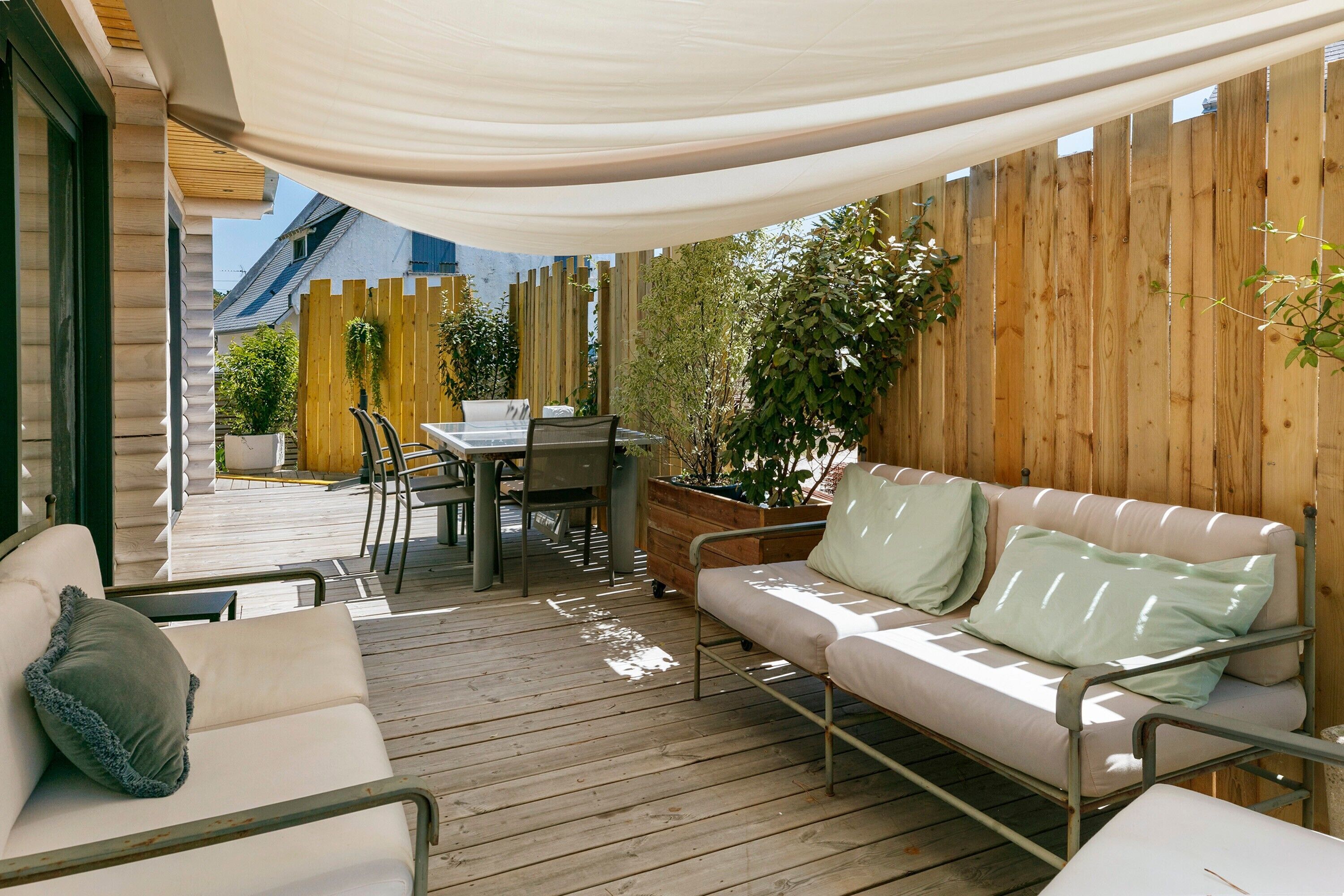 Een overdekt terras met een houten terras, een witte bank, een eettafel met stoelen en een houten omheining.
