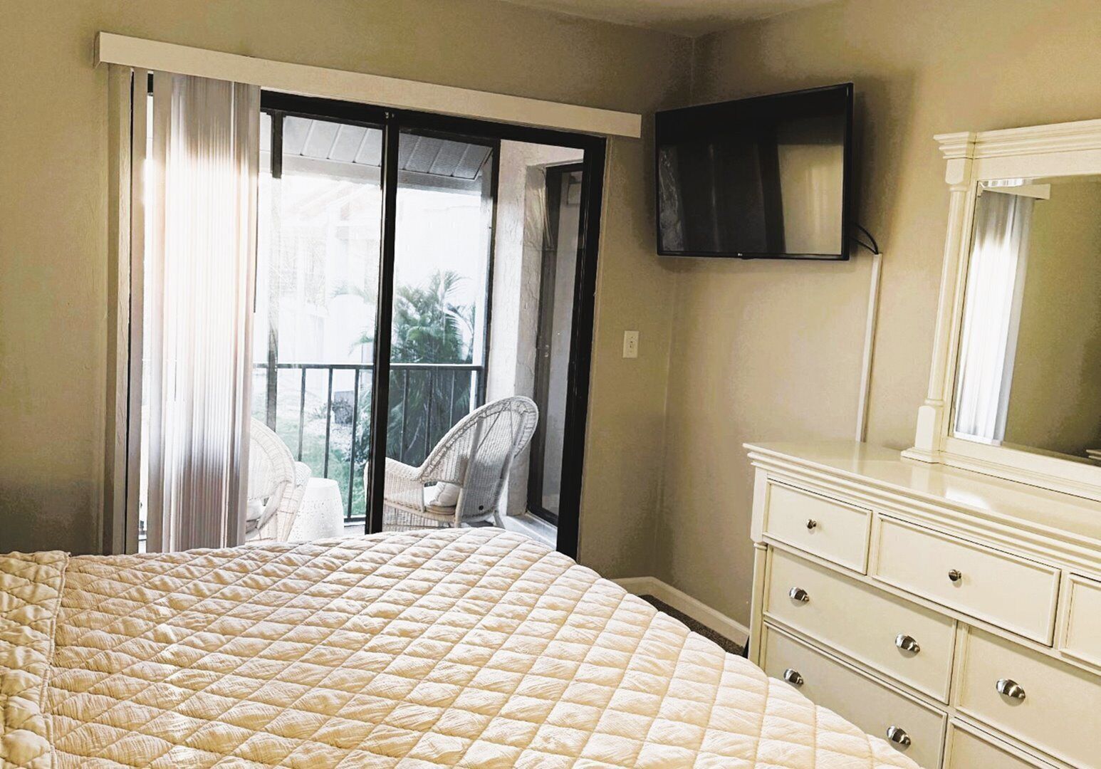 A bedroom with a bed, a chair, a dresser, a TV, and a balcony with curtains.