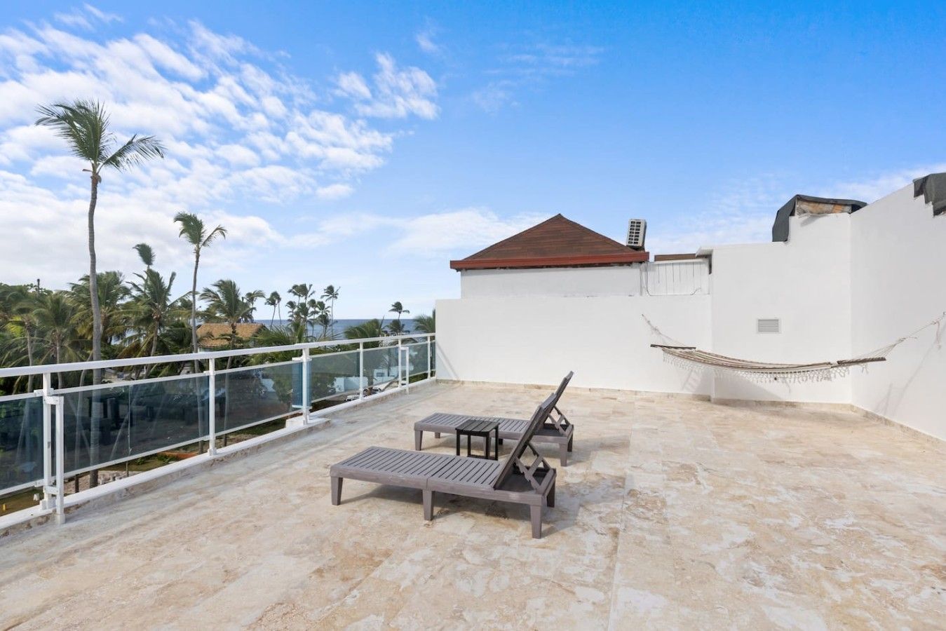 A rooftop terrace with a white wall, a red roof, and a hammock. There are two wooden lounge chairs and a table.