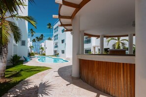 A resort with a pool, palm trees, and a covered area with wooden accents. - Beachfront Penthouse | Electricity Included (Punta Cana)