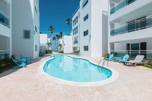 Ein Poolbereich mit einem klaren, blauen Pool, umgeben von weißen Gebäuden und mehreren blauen Stühlen.