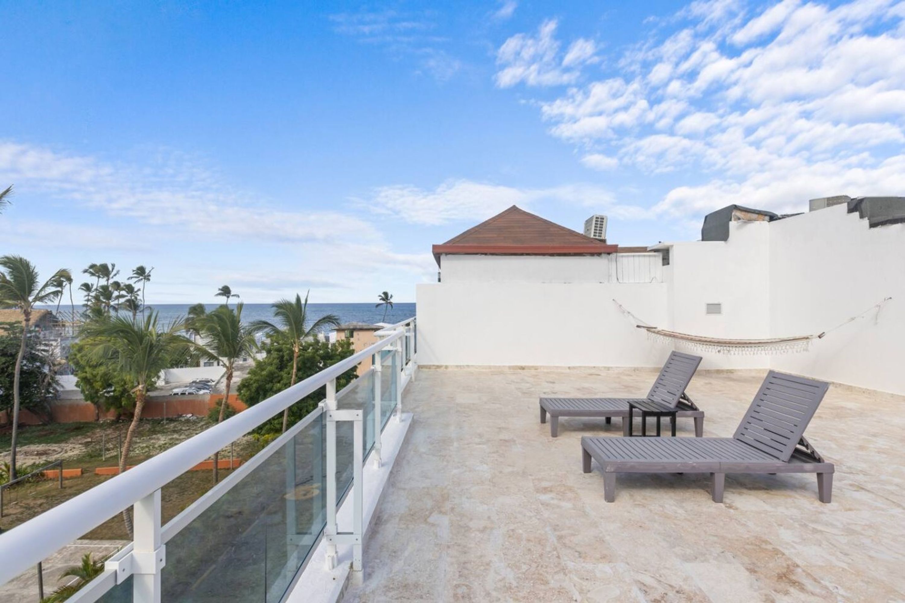 A rooftop terrace with a white wall, a red roof, and a view of the sea. There are two lounge chairs and a hammock.