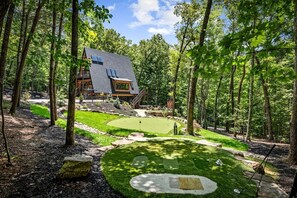 A modern house surrounded by a forest with a well-maintained lawn and a small pond. - Luxe A-Frame | Golf · Hot Tub · National Forest (Morganton)