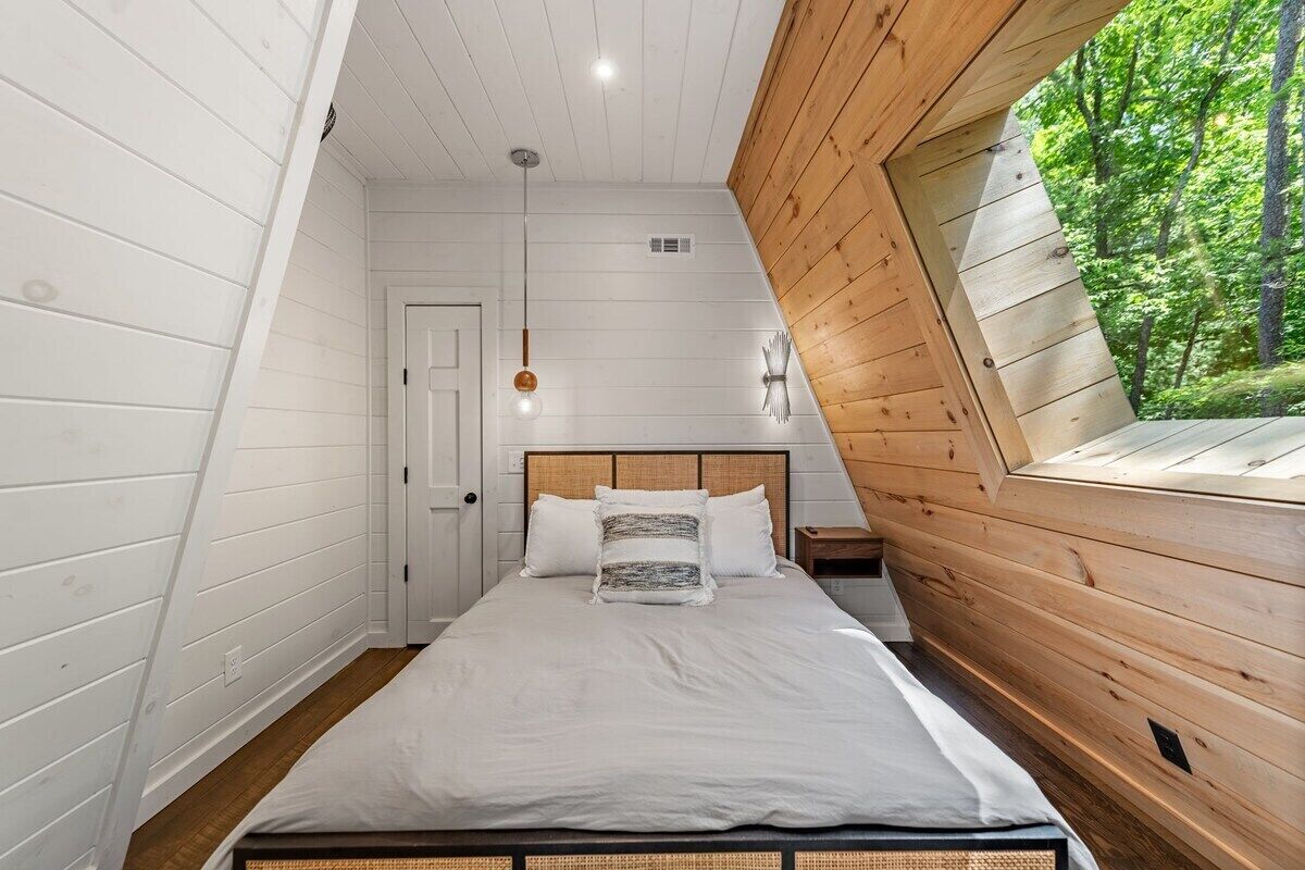 Ein Schlafzimmer mit schrägen Wänden, einem Bett mit weißer Bettwäsche, einem Holz-Kopfteil und einem Fenster mit Blick auf Grünflächen.