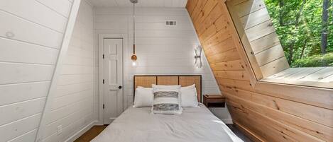 A bedroom with a sloped ceiling, a bed with white bedding, a wooden headboard, and a window overlooking greenery.