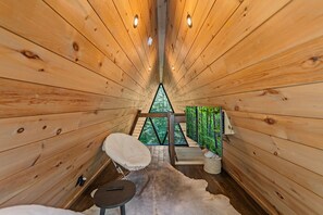 Ein Holzhütten-Innenraum mit schrägverdeckter Decke, einer kleinen Sitzecke und Blick durch ein dreieckiges Fenster.