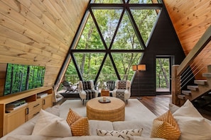 A modern interior with a large triangular window, wooden walls, a sofa set, and a staircase. - Luxe A-Frame | Golf · Hot Tub · National Forest (Morganton)