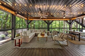 A covered outdoor seating area with a wooden floor, a sofa, chairs, a table, and a ceiling fan. - Luxe A-Frame | Golf · Hot Tub · National Forest (Morganton)