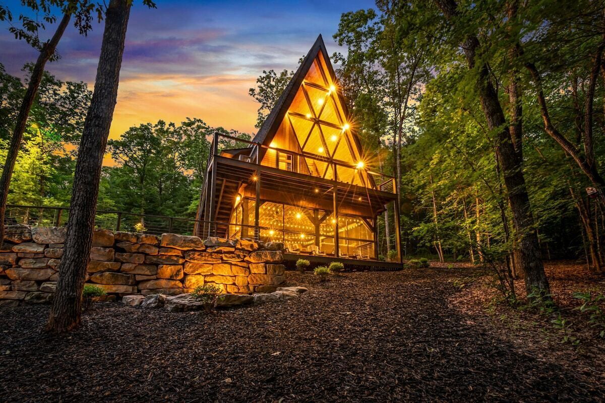 Una cabaña moderna de madera con forma triangular, rodeada de árboles, con un muro de piedra y un interior bien iluminado.