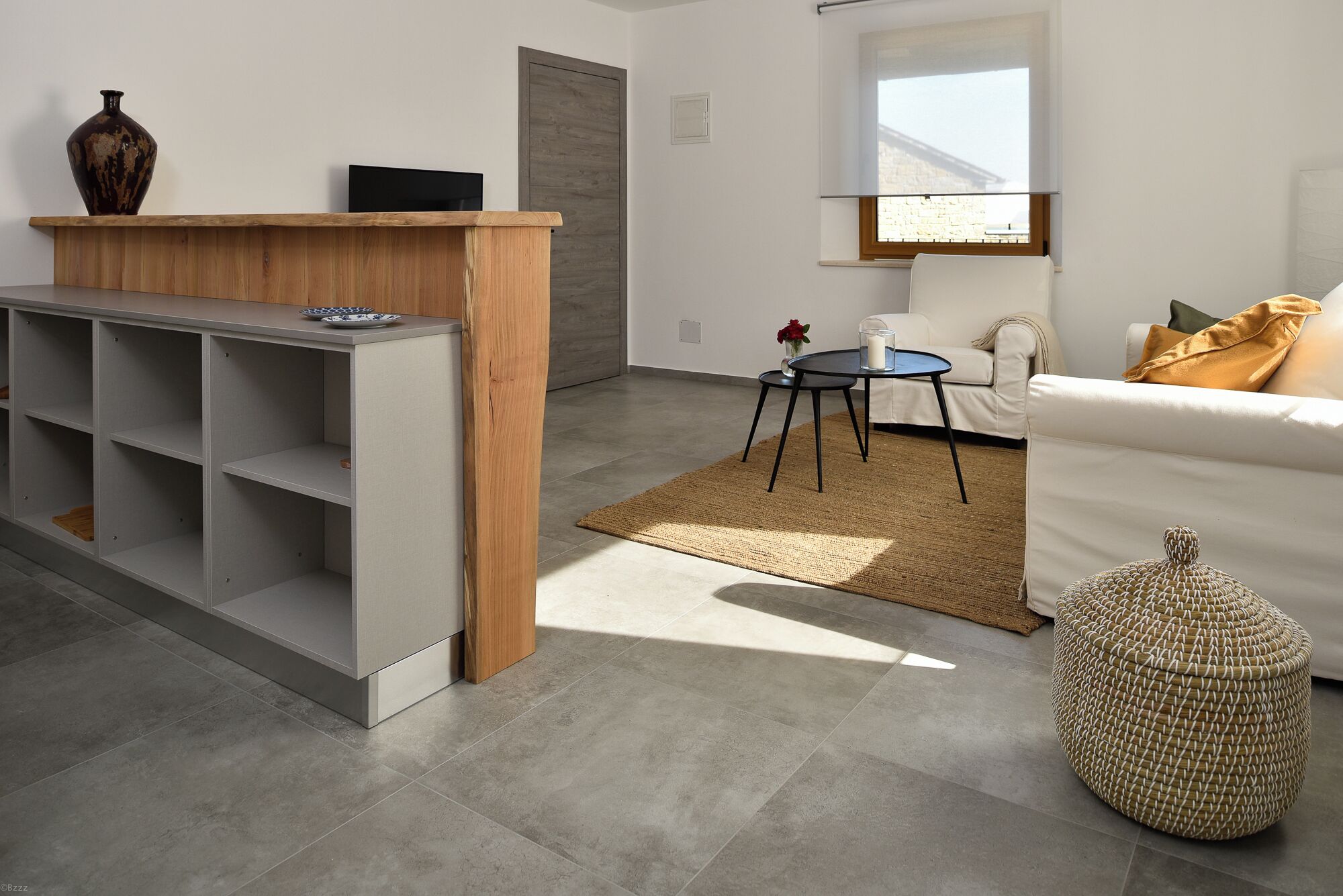 Ein modernes Wohnzimmer mit einem hölzernen Empfangstresen, einem weißen Sofa, einem runden gewebten Pouf und einem kleinen Beistelltisch mit einer Blumenvase.