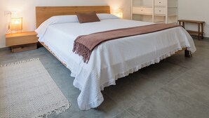 Een net opgemaakt bed met een houten hoofdbord, een witte deken en een bruine plaid. Er staan nachtlampjes, een klein houten nachtkastje en een wit opbergmeubel op de achtergrond.