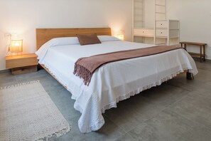 Ein ordentlich bezogenes Bett mit einem hölzernen Kopfteil, einer weißen Decke und einer braunen Tagesdecke. Im Hintergrund stehen Nachttischlampen, ein kleiner Nachttisch aus Holz und eine weiße Kommode.