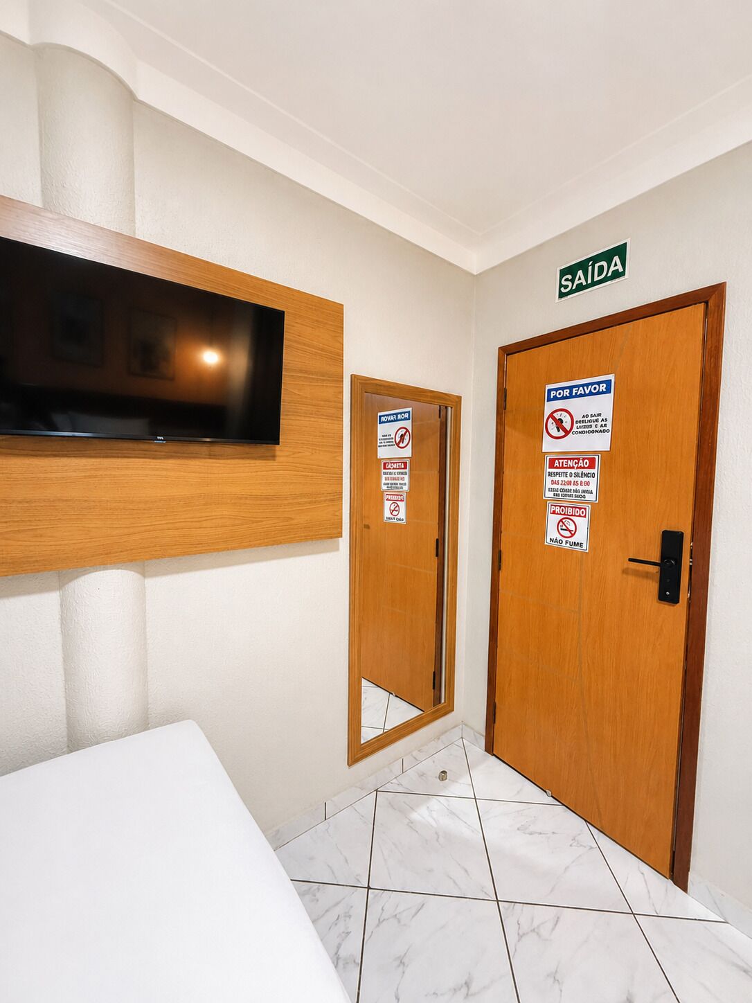 Ein Hotelzimmer mit an der Wand befestigtem Fernseher, einer Holztür mit mehreren Schildern und einem gefliesten Boden.