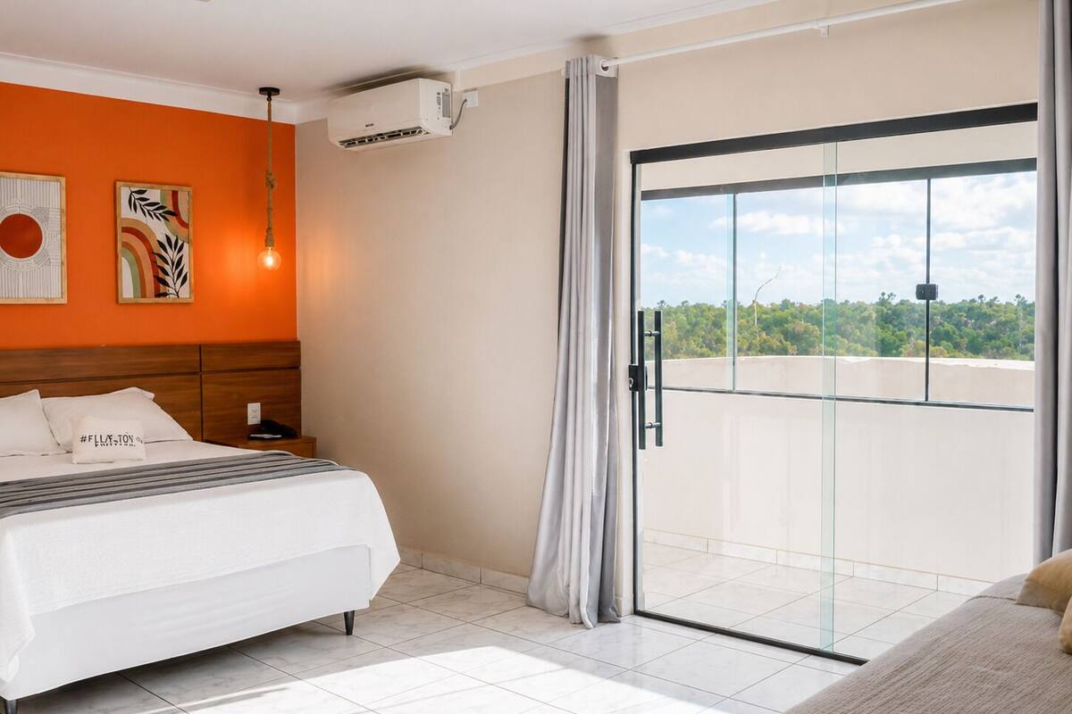 A hotel room with a bed, a nightstand, an air conditioning unit, a window with a view, and a curtain.