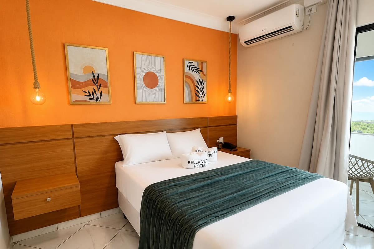 A hotel room with a bed, a nightstand, a wall-mounted air conditioner, and artwork.