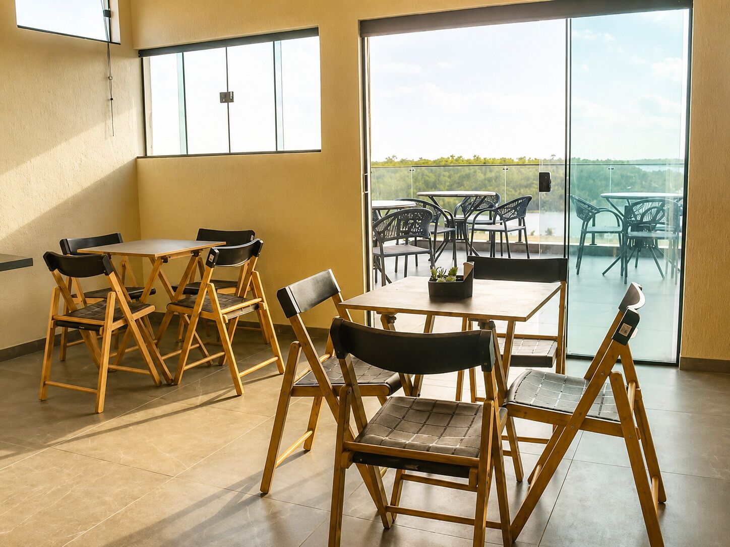 A dining area with foldable chairs and tables, large windows, and a view of outdoor seating.