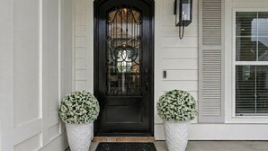 Una puerta frontal negra con un panel de vidrio decorativo, flanqueada por dos macetas blancas con follaje verde, y un farol negro en la pared a la derecha.