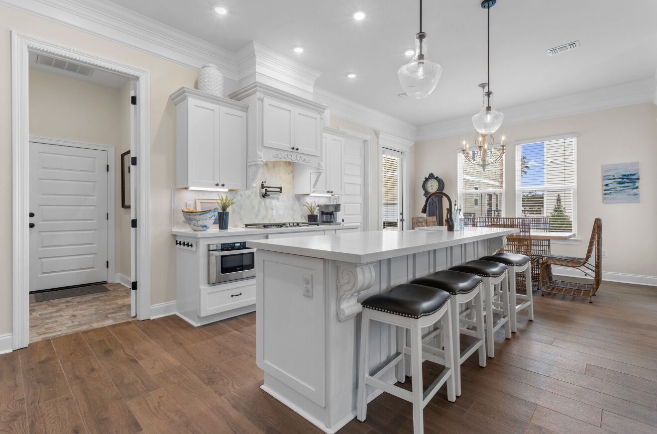 A kitchen with white cabinetry, a central island with bar stools, a dining area with a wicker chair, and a clock on the wall.