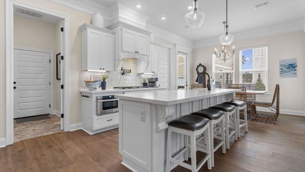 Una cocina con armarios blancos, una isla central con taburetes de bar, un comedor con una silla de mimbre y un reloj en la pared.