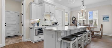 A kitchen with white cabinetry, a central island with bar stools, a dining area with a wicker chair, and a clock on the wall.
