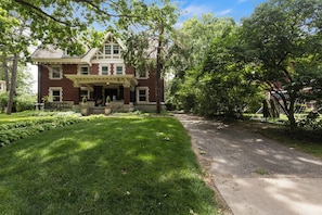 A large brick house with a front porch, surrounded by lush greenery and trees.
