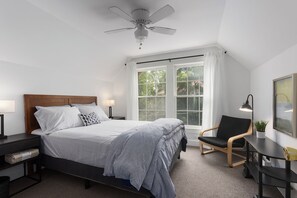 A bedroom with a bed, bedside tables, a chair, a desk, a ceiling fan, and a window with curtains.