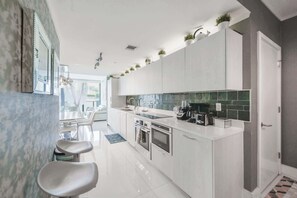 Eine moderne Küche mit weißen Schränken, einer schwarzen Arbeitsplatte und einer grünen Rückwand. Zwei Barhocker und ein Esstisch mit Stühlen sind vorhanden.
