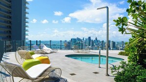 En pool på taket med solstolar, fri utsikt över stadens silhuett och en hög byggnad i bakgrunden.