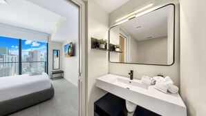 Et moderne hotelværelse med en stor seng, et badeværelse med hvid vask og spejl, og udsigt over byen.