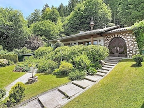 A traditional building with a stone facade, a wooden staircase leading to a door with a leaf design, and a well-maintained garden with various shrubs and a lawn. - Lovely apartment for 3 guests with WIFI, TV, patio and pets allowed (Schiefling Am See)