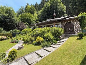 Ein traditionelles Gebäude mit einer Steinfassade, einer Holztreppe, die zu einer verzierten Eingangstür führt, und einem gepflegten Garten mit verschiedenen Sträuchern und einer Rasenfläche.