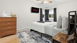 Een slaapkamer met een bed, een houten dressoir, een plafondventilator, een aan de muur bevestigde televisie en een raam met gordijnen.