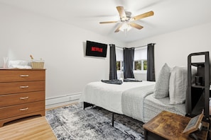 Une chambre à coucher comprenant un lit, une commode en bois, un ventilateur de plafond, un téléviseur fixé au mur et une fenêtre avec des rideaux.