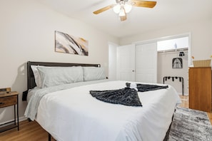 Un dormitorio con cama, mesita de noche, ventilador de techo, una puerta y un estante en la pared.