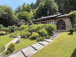 Ein traditionelles Gebäude mit einer Steinfassade, einer Holztreppe, die zu einer verzierten Eingangstür führt, und einem gepflegten Garten mit verschiedenen Sträuchern und einer Rasenfläche.