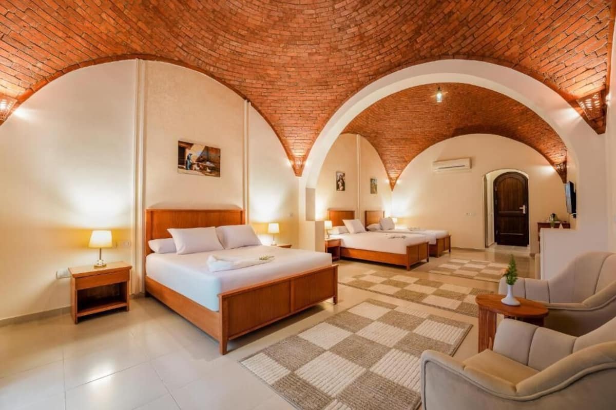 Una habitación de hotel con techo de ladrillo, nichos abovedados, una cama grande, una cama más pequeña, un sofá y un televisor.
