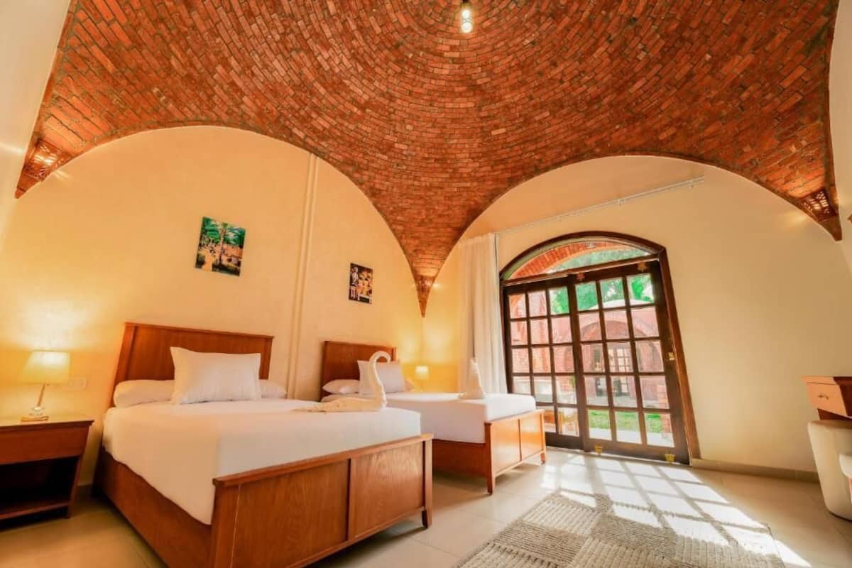 Una habitación con techo de ladrillo, dos camas, un escritorio y un ventanal grande.