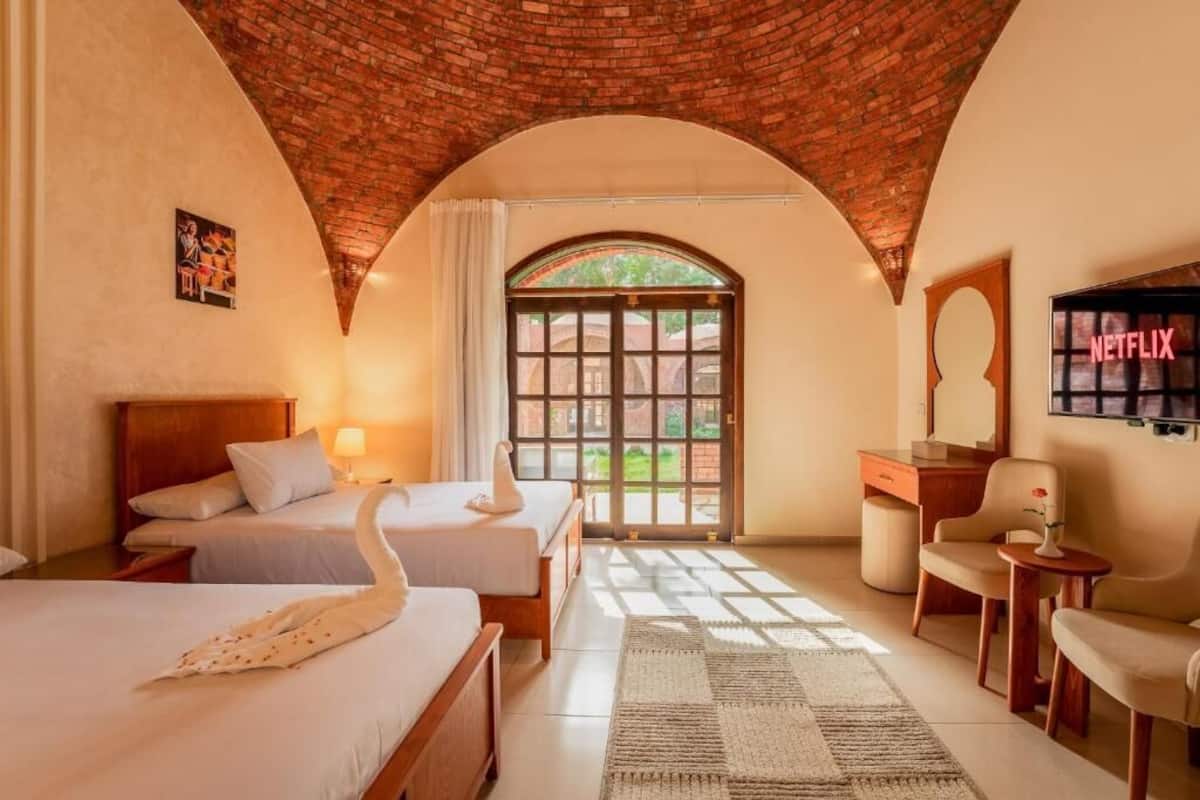 Habitación de hotel con dos camas, un escritorio, una silla, un televisor y un amplio ventanal de arco.