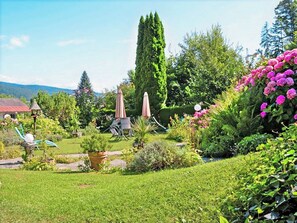 Ein üppiger Garten mit verschiedenen Pflanzen und Blumen, ein hoher Baum und Gartenmöbel.