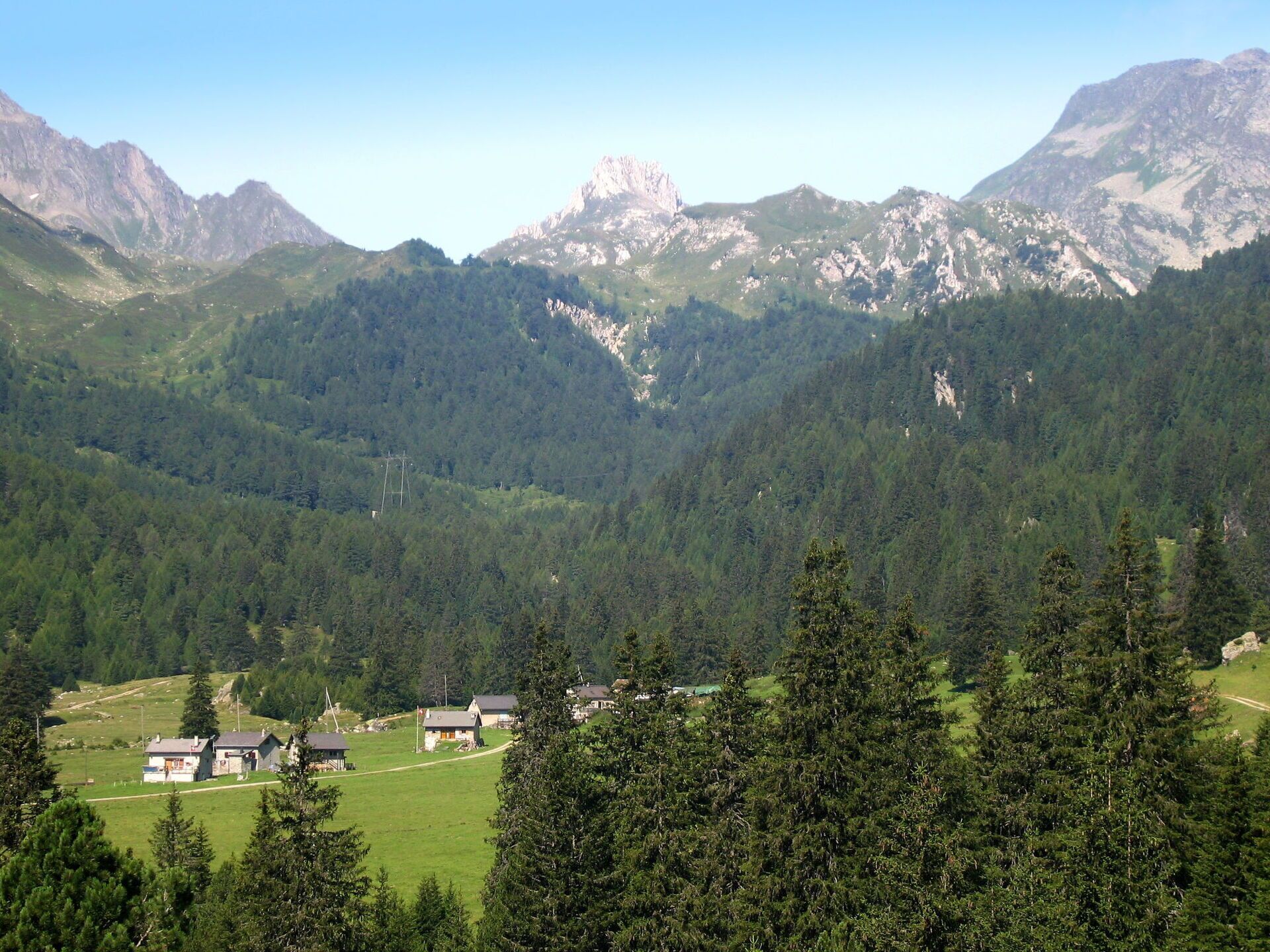 Eine gebirgige Landschaft mit einer kleinen Siedlung, mehreren Gebäuden, dichten Wäldern und felsigen Gipfeln.