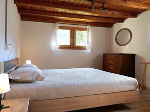 Ein Schlafzimmer mit Holzbalkendecke, einem Bett mit weißer Bettdecke, einem Schminktisch und einem Fenster mit Spitzenvorhängen.