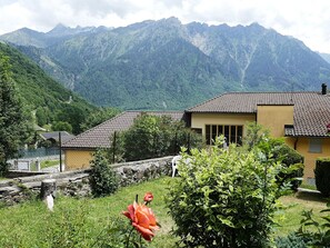 Ein Haus mit Ziegeldach, umgeben von Grün und Bergen im Hintergrund.
