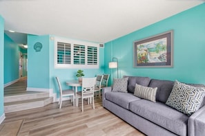 A living room with a grey sofa, a dining table with chairs, a window with shutters, and a painting on the wall. - Beach Cove Resort 1014 - Oceanfront Condo with Direct Beach Access (North Myrtle Beach)