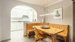 Uma área de refeições com um banco de madeira, uma mesa e uma tigela. Cozinha ao fundo com armários brancos e uma janela.
