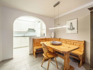 Ein Essbereich mit einer hölzernen Bank, einem Tisch und einer Schüssel. Im Hintergrund ist eine Küche mit weißen Schränken und einem Fenster zu sehen.