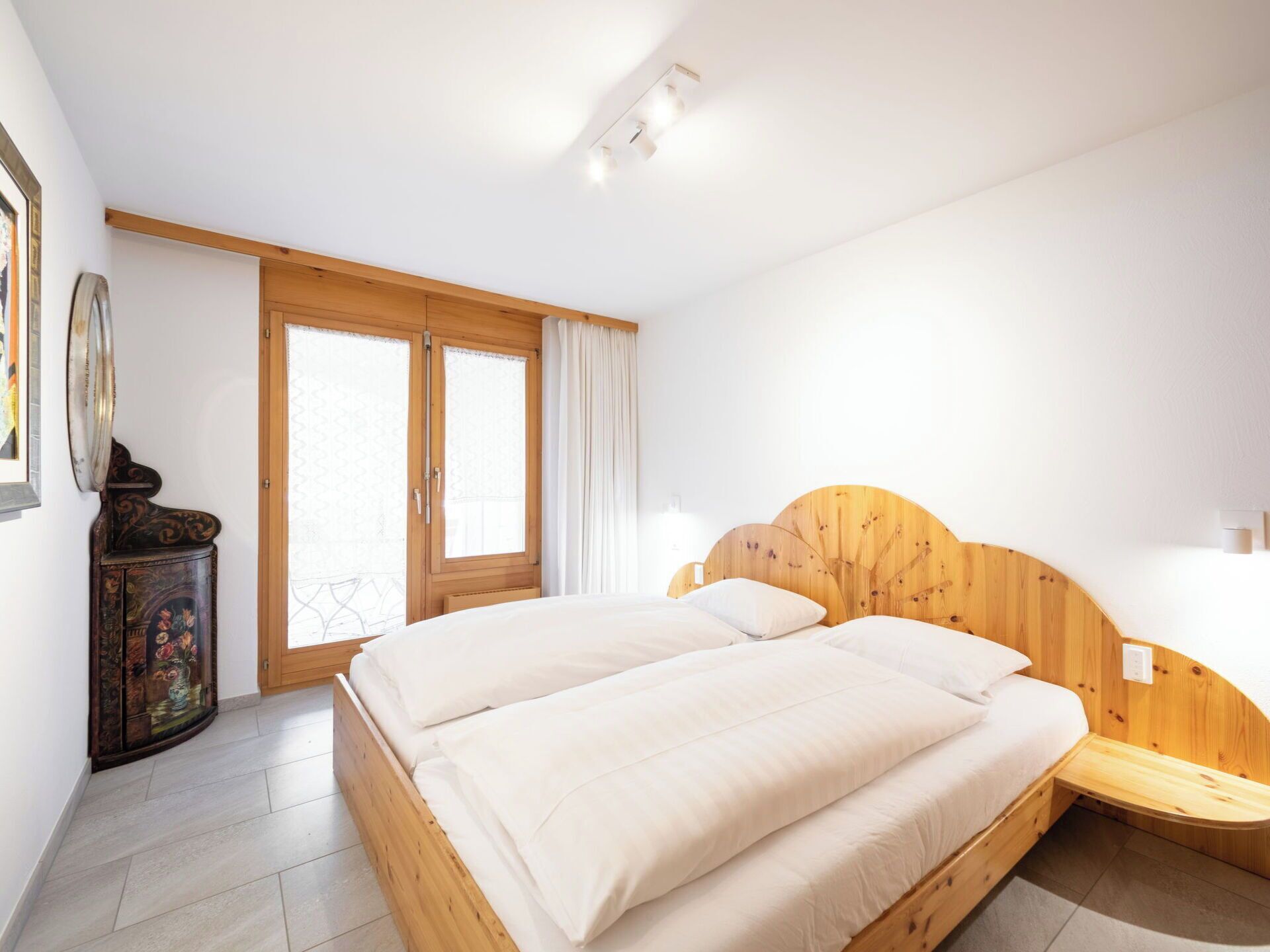 Una camera da letto con un letto di legno, un comodino in legno, una cassettiera e un'ampia finestra con tende.