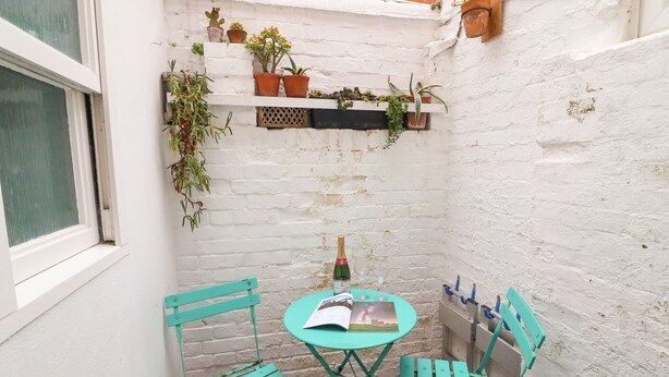 A small outdoor seating area with a table and two chairs, a bottle of champagne, and potted plants on a white brick wall. - Pixie Cottage (Weymouth)